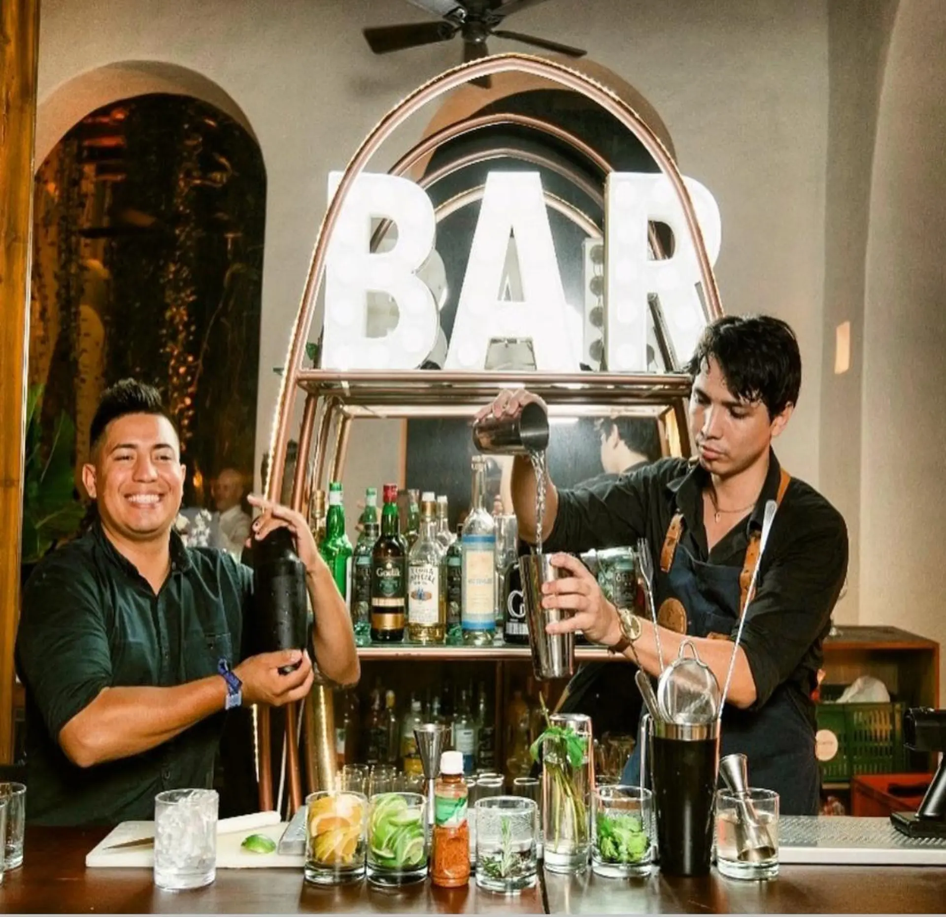 Dos bartenders de Stage preparando cócteles en una barra iluminada con letras BAR